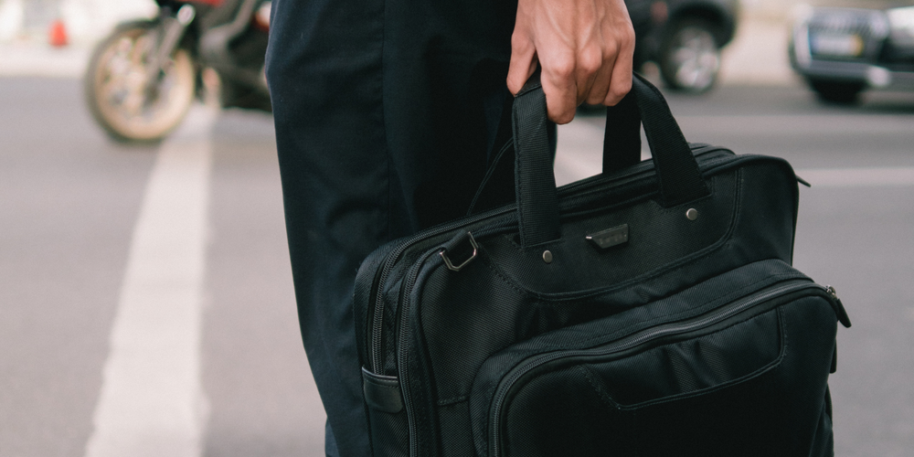 Mann in schwarzer Kleidung, hält schwarze Laptoptasche am Griff, Straße mit Motorrad im Hintergrund