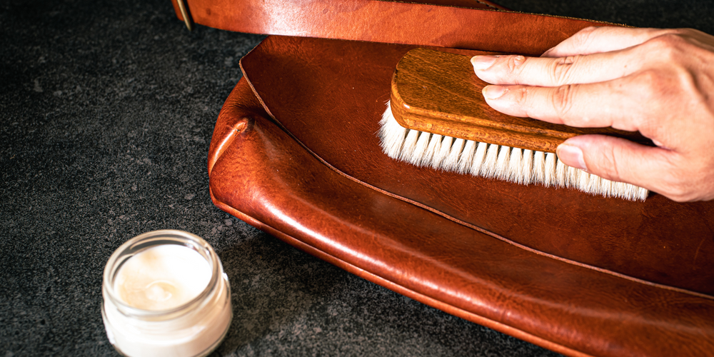Hand mit Bürste, Lederpflege auf brauner Ledertasche, Pflegemittel im Glas, Lederpflege, Accessoires