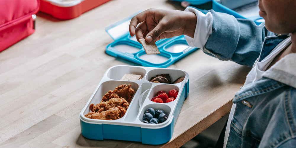 Lunchbox, auf Holztisch, verschiedene Fächer mit Obst und Snacks, Kind mit Jeansjacke, greift nach Snack