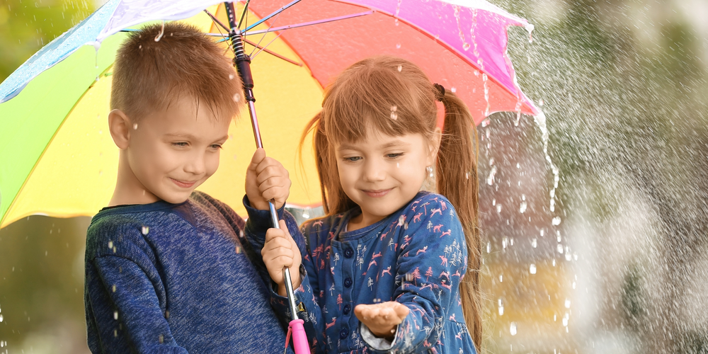 Regenschirme für Kinder, zwei Kinder unter buntem Schirm, lachend, Regen fällt, Mädchen hält Hand nach vorn