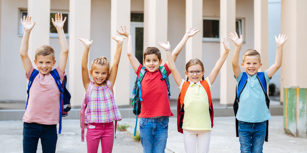 Schule, fünf Kinder mit bunten Schulrucksäcken, halten die Hände in die Luft, stehen vor einem Gebäude, fröhliche Stimmung, Schulbeginn