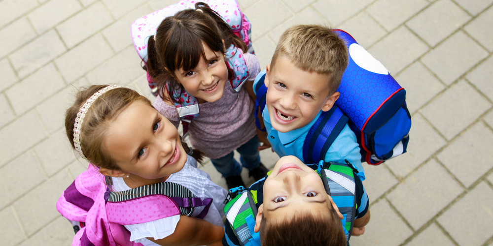 Schulranzen-Sets, vier Kinder mit Schulranzen, von oben fotografiert, schauen lachend in die Kamera, gepflasterter Boden, bunte Ranzen