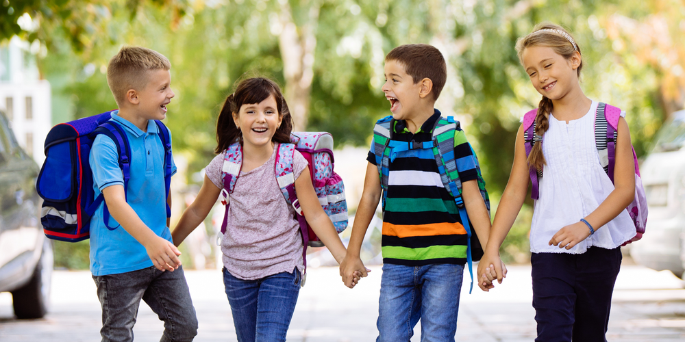 Schulranzen, vier Kinder mit bunten Schulranzen, halten sich an den Händen, gehen auf dem Gehweg, fröhliche Stimmung, Schulanfang