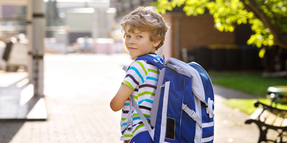 Schulranzen für Jungs, Junge mit blauem Schulranzen, blickt über die Schulter, trägt gestreiftes T-Shirt, helle Straße, sonniger Tag