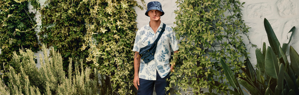 Camel Active, Mann mit Bucket Hat, lässiges Sommeroutfit, Umhängetasche quer getragen, grüner Pflanzenhintergrund, Outdoor- und Freizeitlook, urbanes Natursetting