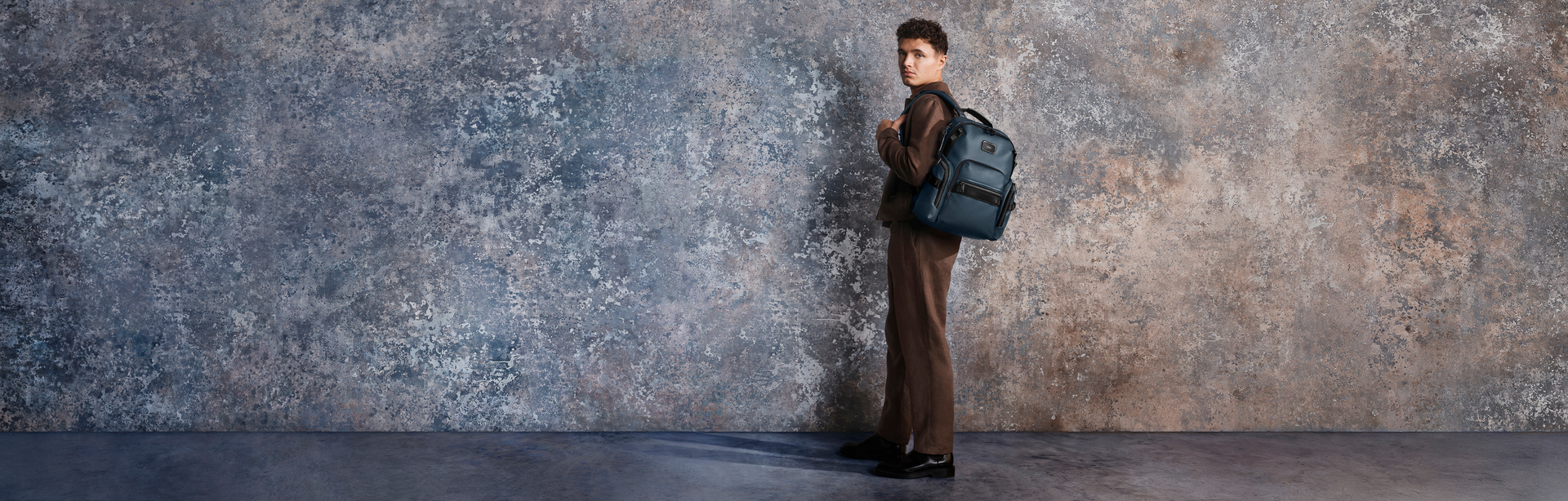 TUMI, McLaren Formel-1-Fahrers Lando Norris, Rucksack, Mann steht seitlich mit schwarzem Rucksack, braunes Outfit, minimalistisches Studio-Setting, Betonwand-Hintergrund, moderner Business-Look, Premium-Accessoire