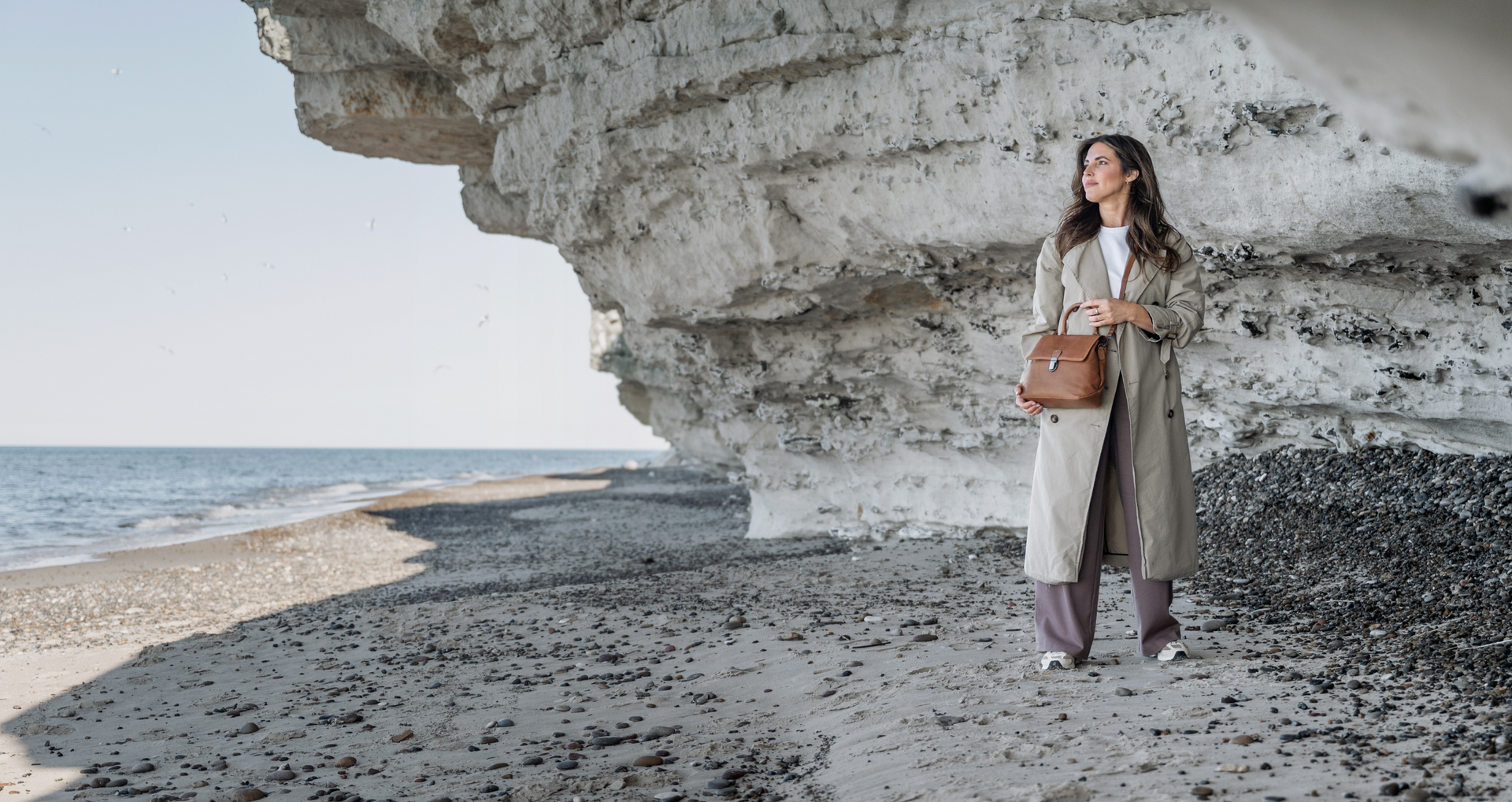 The Chesterfield Brand, Ledertasche, cognacbraun, Trenchcoat, Person im Mantel, Steinwand, Kiesboden, herbstlich, zeitlos, hochwertig, Vintage-Look