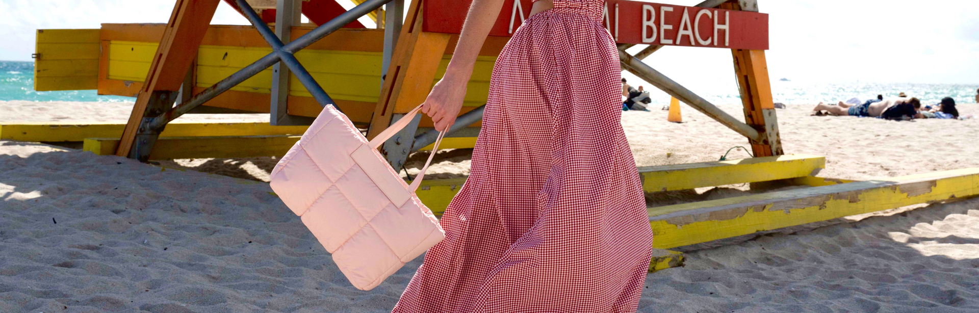 VeeCollective, rosa Handtasche, gestepptes Design, Frau im pinken Kleid, Sandstrand, Freizeitlook, moderne Mode, Outdoor-Szene, stilvoll, sommerlich