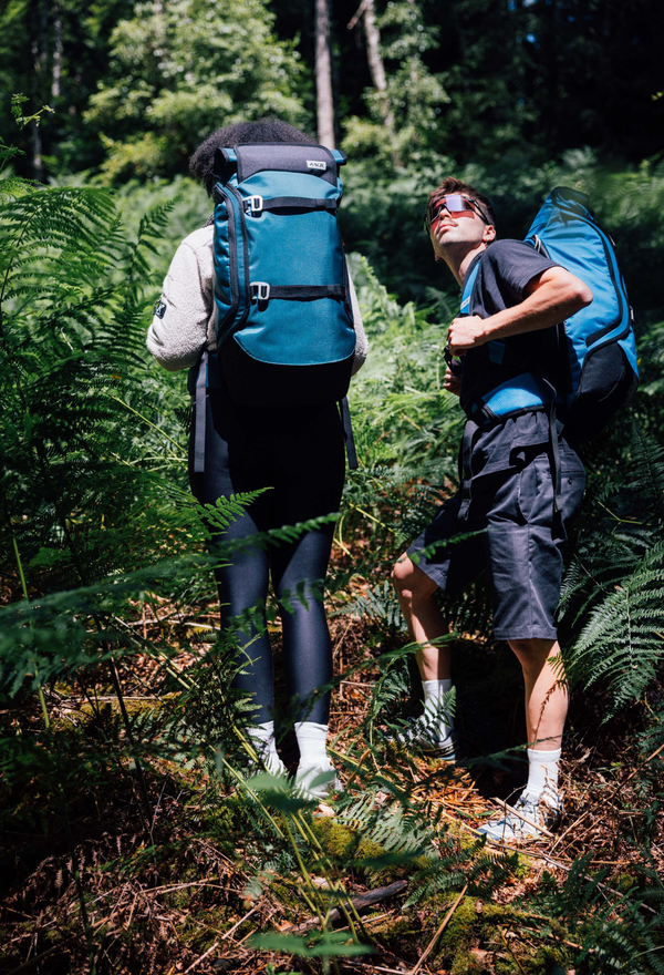 Aevor, junge Person sitzt auf Wiese, mehrere Outdoor-Rucksäcke gestapelt, schwarze und blaue Rucksäcke, entspannte Pose, nachhaltige Reiserucksäcke, Outdoor- und Abenteuer-Setting
